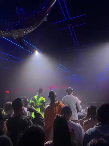 The video shows a dark, indoor venue, likely a club or concert space, filled with a crowd of people. On a raised platform or stage, several individuals are visible, with at least two appearing to be operating DJ equipment. Bright spotlights illuminate the stage from above, and blue neon-like lights crisscross the ceiling, casting geometric patterns. A large, reflective object, possibly a disco ball, hangs from the ceiling. Red light dots are also visible in the background. The people in the crowd are seen from behind, generally facing the stage, suggesting they are watching a performance or listening to music. The overall atmosphere appears energetic and engaging, typical of a live music event. The video briefly pans down to show more of the crowd and their upper bodies.