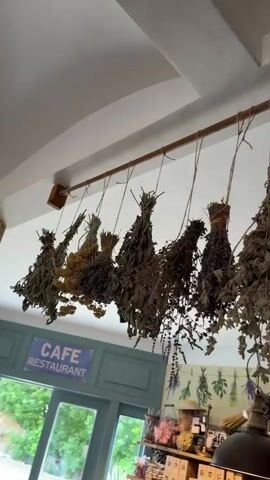 A low-angle shot shows bundles of dried herbs and flowers hanging from a wooden rod attached to a white ceiling. The bundles are suspended by twine and vary in size and color, with some appearing to be lavender and others dried grasses. Below the hanging herbs, a blue wall is visible with a sign that reads "CAFE RESTAURANT." Further down, a wooden shelf displays various items, including bottles and boxes. The lighting suggests an indoor environment, possibly a shop or cafe.