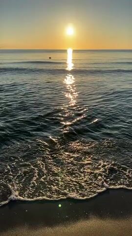 This is a video of a sunrise over the ocean. The sun is a bright orb in the sky, casting a shimmering reflection on the water. Small waves are lapping at a sandy shore in the foreground. The sky is a clear, soft yellow.