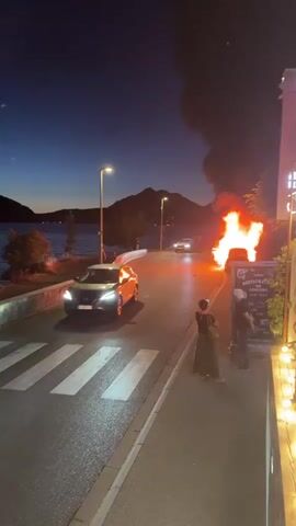 The video captures a scene at dusk or night with a dark blue sky. A road runs diagonally across the frame. On the left side of the road, there is a body of water and a stone wall. Further up the road, a car is seen driving away from the camera. On the right side of the road, a car is engulfed in flames, emitting a large plume of black smoke that rises into the sky. Two individuals stand on the sidewalk near the burning car, observing the event. One person is wearing a dark green dress and holding a bag, while the other is wearing a light-colored shirt and dark pants. A sign with text and images is visible next to them. The road has a pedestrian crossing marked with white stripes. Streetlights illuminate the scene, casting light on the road and the surroundings. In the background, hills or mountains are silhouetted against the sky. The overall atmosphere suggests a dramatic and potentially dangerous event.
