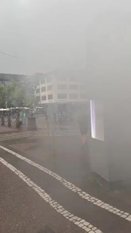 The video shows a street view of a city on a cloudy day. Several individuals are walking along the sidewalk, some holding umbrellas, and a bus is seen in the background. A tall, red brick monument or column stands on the left side of the frame. On the right, a gray informational sign with a green top displaying the number "6" and text is visible.