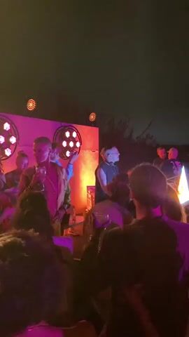 A group of people are gathered outdoors at night. The scene is illuminated by stage lights with pink and yellow hues, and some spotlights. In the background, a band or performers are on a stage. Some audience members are visible in the foreground, many with their backs to the camera, some holding drinks. The overall atmosphere appears to be that of an outdoor concert or performance.
