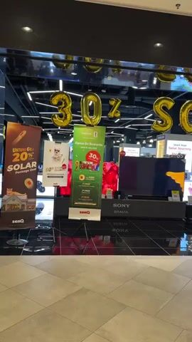 The video shows the storefront of a Grand senQ electronics store. Large gold balloons spelling out "30% OFF" are hanging above the entrance. Several promotional banners are displayed outside the store, advertising deals on solar packages and other items. A large television screen displays a bright, abstract orange and yellow image. The store is well-lit, with a modern, sleek design. The floor is tiled and reflective, showing the reflections of the store's interior and the promotional displays. A portion of the store's interior is visible, including shelves stocked with electronics and some staff members.