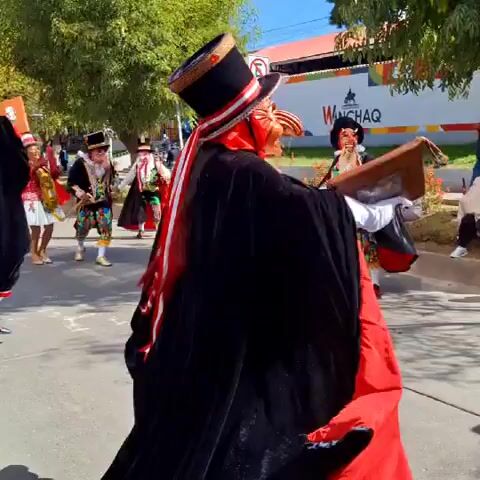 A group of people in colorful costumes and masks are participating in a parade or procession on a paved street under a clear blue sky. The foreground features a performer wearing an elaborate mask with a prominent nose and a black top hat. They are dressed in a black cape lined with red, a white ruffled shirt, and a red vest adorned with gold and white embroidery. They are holding a small wooden frame in their right hand and their left arm is bent as if holding something hidden within the cape. Behind them, other performers in similar attire, some holding whips and other props, are also visible, moving along the street. Spectators are seated and standing on the side of the road, observing the event. Trees line the street, and buildings are visible in the background, one of which has a sign with a stylized logo and the text "WANCHAQ". The overall atmosphere appears festive.