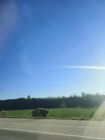 The video shows a view from inside a vehicle, looking out at a bright blue sky with the sun shining. A green field with trees lining the horizon is visible. A road is in the foreground. The video appears to be taken while the vehicle is moving.