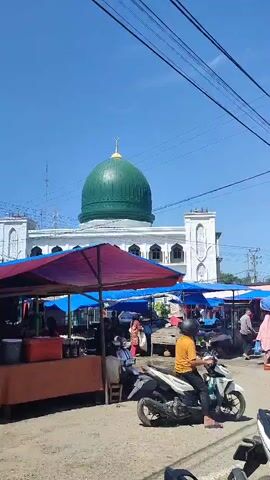 The video shows a street scene in front of a mosque with a green dome. There are several market stalls set up along the street, with vendors selling various goods under colorful awnings. People are seen walking around and browsing the stalls. A few motorcycles are also visible, with some people riding on them. The sky is clear and blue, indicating a sunny day.