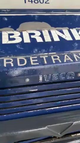 The video shows a Brink's Transport armored truck parked on a wet, cobblestone street in a city. The truck is blue and white, with the words "BRINK'S TRANSPORT" printed on its side. The street is lined with parked cars, trees, and buildings. A Moroccan flag is visible on a building in the background. It is raining.