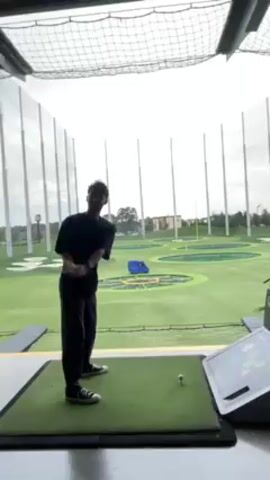 The video shows a person, likely male, in a Topgolf facility swinging a golf club. He is wearing a black shirt, black pants, and black Converse sneakers. He’s standing on a green mat, and a golf ball is teed up in front of him. Behind him is the open-air driving range with targets on the ground. A touchscreen panel is visible to his right. He takes a swing at the ball. The netting and support structures of the Topgolf facility are visible in the background.