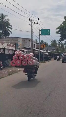 A black motorcycle laden with large rolls of what appear to be carpeting is driving on a paved road in a populated area with overhead power lines, road signs, and various commercial buildings. The rolls are wrapped in clear plastic and have a red, web-like pattern visible at their ends. The motorcycle is moving away from the camera, and there are other vehicles and motorcycles on the road in the same direction. The weather is overcast, and trees can be seen in the background.
