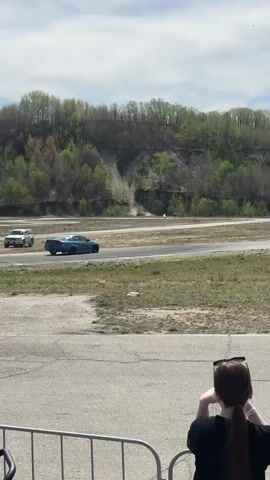 The video shows a light blue, four-door sedan drifting on a paved racetrack. The car is drifting to the left of the frame, leaving trails of smoke. The video is taken from behind a fence, with spectators watching the car perform. The background includes trees, hills, and the sky is mostly clear with some clouds. The racetrack is surrounded by grass and gravel. There are utility poles in the distance. The spectators appear to be mostly young adults.