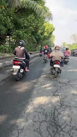 A group of motorcyclists are riding down a paved road. The road is lined with trees and a curb. The sky is clear. The motorcyclists are wearing helmets and casual clothing. The motorcyclists are riding in single file. The video is shot from behind the motorcyclists.