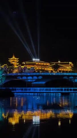 At night, a dark body of water reflects a brightly lit, traditional Chinese building. A pagoda sits to the left of the building. The building is covered in lights, with white lights shining up into the night sky. A bridge sits between the viewer and the building, with water flowing over a short waterfall into the river in front of the bridge. The buildings are reflected in the water, creating a beautiful symmetrical image.