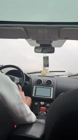 The video shows the interior of a car from the perspective of a passenger, looking towards the driver's seat and the windshield. The windshield wipers are intermittently clearing rain from the glass, indicating it is raining outside. The driver's hands are visible on the steering wheel. The car has a sunroof, which is open. A rearview mirror hangs from the windshield, with an air freshener dangling from it. The car's dashboard is visible, including the infotainment system, air vents, and a bottle of water with an orange cap. Another passenger's shoulder and head are partially visible in the right side of the frame. The overall environment appears to be a gray, overcast day due to the rain.