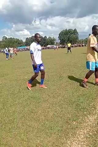 A video captures a soccer game on a grassy field under a cloudy sky. Two soccer players, one in a white and blue uniform and the other in a tan and blue uniform, walk along the sideline. The tan and blue uniformed player then enters the field of play as the play moves to the opposite side of the field. Other players wearing the same tan and blue uniform can be seen running downfield. A crowd of spectators line the edge of the field. Trees and a building are in the background.