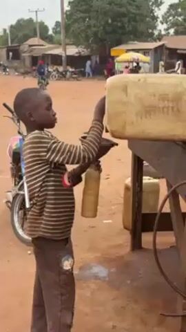 A young boy, probably in his early teens, is transferring liquid from a large plastic container to a smaller one, using a funnel. He is standing next to a metal table, on a dirt lot. The large container is resting on the table.  There are motorcycles parked nearby. Other people and buildings are visible in the background. The scene appears to be in a village or small town. The weather appears overcast.