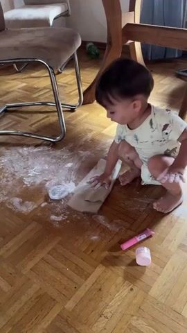 A toddler kneels on a wooden floor, surrounded by a white powdery substance that has been spilled from a small container. The toddler, wearing a light-colored onesie, is using a piece of paper to wipe the powder. A pink tube and a small pink container are also on the floor near the toddler. In the background, a wooden chair and table are visible, as well as another chair with a brown seat.