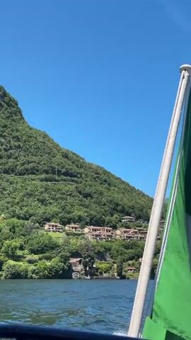 The video shows a body of water with a green flag attached to a pole on the right side. In the background, there is a lush green hillside with several buildings nestled into the landscape. The sky is clear and blue. The water appears calm with some ripples, suggesting movement, possibly from a boat. The camera angle indicates the viewer is on a boat looking towards the shore.