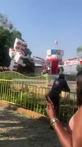 The video shows an amusement park ride called "Energy Storm" in operation. The ride consists of a central tower with rotating arms that lift and spin individual passenger cars. The ride's structure is primarily white with red flame accents. A small flag flutters on top of the tower. A yellow metal fence surrounds the ride. Several people stand watching, including a person with long red fingernails holding a phone and a security guard in a black uniform. The sky is clear and blue, suggesting a sunny day. The background includes trees and a glimpse of other park structures.