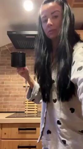 A woman with long dark hair, wearing gray pajamas with black hearts, is in a kitchen. She holds a black mug and stirs the contents with a small red spoon. The kitchen features light wood cabinets, a brick backsplash, and a black range hood. A wooden drying rack sits on the counter behind her.