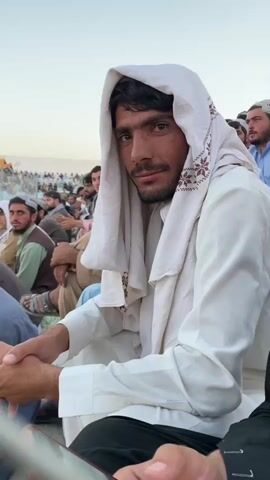The video shows a group of men sitting together outdoors, likely at an event. The focus is on a man in the foreground wearing a white headscarf with embroidered trim. He looks at the camera, then smiles and looks away. Other men are visible in the background, some wearing traditional hats. The environment appears to be dusty, with a large crowd and structures in the distance. The lighting suggests it is daytime, possibly late afternoon.