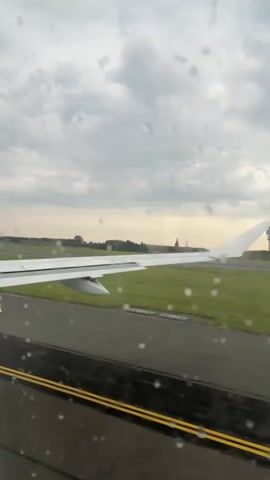 The video begins with a view out of an airplane window. Raindrops are visible on the windowpane, distorting the view of the exterior. The camera pans slightly to the right, revealing the white wing of the airplane. Beyond the wing, a green grassy field is visible, bordered by a tarmac runway with yellow and black stripes. In the distance, a structure that appears to be an airport control tower can be seen. The sky above is overcast with clouds. The airplane is stationary on the runway, suggesting it is either preparing for takeoff or has just landed.