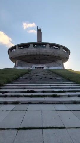 This media file captures a low-angle, ground-level view looking up at the Buzludzha Monument in Shipka, Bulgaria. The monument, a Brutalist structure resembling a flying saucer with a central tower, is situated on a grassy hilltop. A wide, long flight of stone stairs leads up to the monument's entrance, with the lower steps covered by larger paving stones. Green grass covers the slopes on either side of the stairs. The sky above is a pale blue with scattered, wispy white clouds, suggesting it is either early morning or late afternoon. The overall mood is one of solemnity and perhaps a touch of desolation, given the monument's abandoned state, with a sense of grandeur from its imposing architecture. The lighting indicates the sun is low in the sky. There are no people visible in the frame. The monument itself is made of concrete and appears weathered, with some visible graffiti or faded markings on its lower facade. The central tower has a distinctive, almost lighthouse-like structure at its peak.