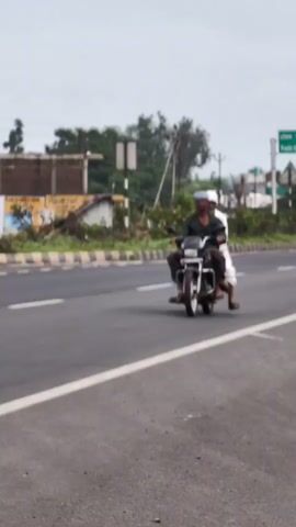 TWO MEN ARE RIDING ON A MOTORCYCLE ON A PAVED ROAD. THE MAN IN FRONT IS DRIVING. THE MAN BEHIND IS SITTING BEHIND THE DRIVER. BOTH MEN ARE WEARING TURBANS. THE MOTORCYCLE IS SILVER. THE ROAD HAS MULTIPLE LANES. THERE ARE TREES AND BUILDINGS IN THE BACKGROUND. THE SKY IS CLOUDY.