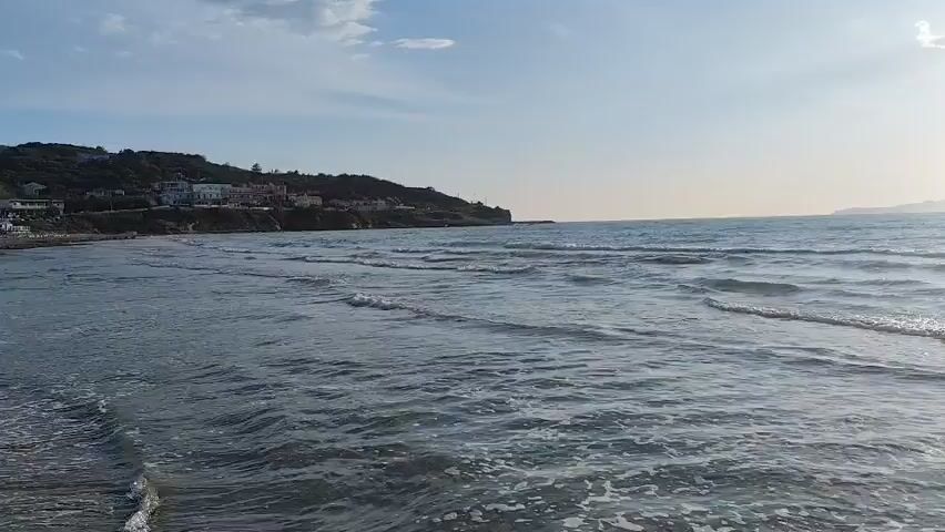 The camera pans across a beach towards the sea. The water is calm with small waves coming into shore. The sun is bright, reflecting off the water. In the distance, a land mass is visible, possibly an island. A hillside with some buildings on it is visible to the left of the frame. The sky is clear with some clouds.