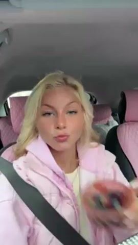 A young woman with blonde hair, wearing a pink jacket, sits in the driver's seat of a car. The car's interior has pink seat covers. She holds a small pink bowl containing blueberries and grapes. She puckers her lips towards the camera, then opens her mouth as if she's about to eat some of the fruit. There is music playing in the background.