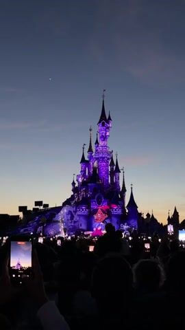 A crowd of people are silhouetted against a purple-lit castle. The castle is adorned with lights and projections of cartoon characters. The sky is a deep twilight blue with a hint of pink. Many people are holding up cell phones to record the spectacle.