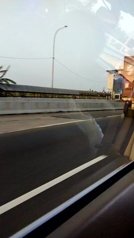 The video shows a view through a window, possibly from a moving vehicle. The scene outside is blurry and consists of a road with white lines, a sidewalk or barrier, and some buildings in the distance. There is also a street light visible. The lighting suggests it is daytime, but the sky appears overcast or hazy. The camera angle is vertical, giving a distorted perspective. There are no people or clear actions discernible in the video.
