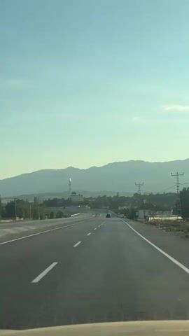 The video captures a view from a vehicle traveling on a highway. The highway is asphalt with white lane markings and shoulders. In the distance, there is a clear blue sky with a faint haze near the horizon. A range of mountains forms the backdrop. On the right side of the highway, there are power lines and utility poles. In the middle distance, a mosque with a green dome and a tall minaret can be seen, situated amongst trees and buildings. Several cars are visible on the highway, and some appear to be passing or merging. The overall impression is of a journey through a landscape with a blend of natural and man-made features.