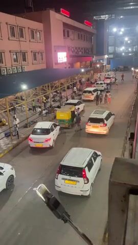 A high-angle shot captures a busy outdoor area at night, likely a train station or bus terminal, with a pink building in the background. A covered walkway with a yellow metal frame and a blue roof runs along the front of the building. Underneath the walkway, numerous people are resting or waiting, some lying on the tiled ground with luggage beside them, others sitting. The area is lit by overhead lights. In front of the walkway, a paved area has several cars and an auto-rickshaw parked. Cars are lined up, suggesting ongoing arrivals or departures. A lamppost is visible in the foreground. The overall impression is one of a transit hub with people waiting during the night.