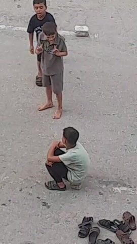 A group of three young boys are on a paved outdoor surface. Two boys stand facing each other in the upper portion of the frame, while a third boy sits in a crouched position in the lower portion. The two standing boys appear to be interacting, possibly playing or arguing, as they gesture towards each other. The boy in the foreground is observing them. There are several pairs of sandals scattered on the ground near the seated boy. The ground is a rough, greyish pavement with some white markings. The lighting suggests it is daytime.