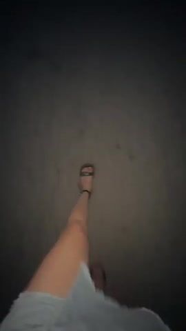 The video displays a first-person perspective of someone walking at night. The scene is dark, suggesting it is nighttime. The ground appears to be asphalt or a similar paved surface, with some scattered debris or marks. The camera is focused on the walker's legs and feet, with a denim skirt or shorts prominently visible in the foreground. The person is wearing sandals. A hand with red nail polish is briefly visible on the left side of the frame. The overall impression is of a casual walk in an urban or semi-urban environment after dark. There is no visible text in the video.