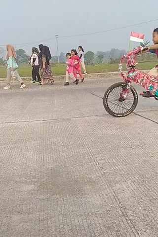 The video shows a young boy on a bicycle performing a stunt. The bicycle is decorated with colorful streamers. The boy is wearing a red shirt and tan shorts. Several other people are visible in the background, some of whom appear to be watching the stunt. The scene takes place on a paved road with a grassy field and trees in the background. The sky is overcast. The video is filmed vertically.