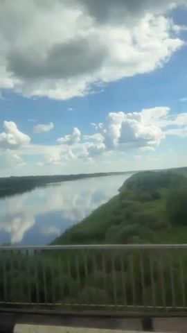 The video captures a wide, calm river viewed from a moving vehicle crossing a bridge. The sky is bright blue with scattered, fluffy white cumulus clouds, which are perfectly mirrored on the still surface of the river. Lush green vegetation, including trees and bushes, lines both banks of the river, stretching into the distance. A metal railing from the bridge is visible in the foreground, framing the lower part of the shot. The scene depicts a serene daytime landscape, likely taken on a sunny day given the clear sky and cloud reflections. The location is in or near Kolomna, Russia, suggesting the river is likely the Oka. No people or visible text are present in the video.