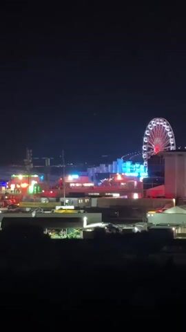 A nighttime aerial view shows a brightly lit amusement park. In the foreground, dark hills are visible. Further back, a Ferris wheel, illuminated with red lights, dominates the scene, its spokes visible against the dark sky. To the left of the Ferris wheel, various carnival rides and stalls are aglow with colorful lights, including green and pink. In the background, a body of water reflects some lights, and a bridge or structure with blue lighting spans across it. The overall impression is of a lively and festive atmosphere.