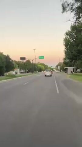 A vehicle travels down an asphalt road during twilight. The sky exhibits hues of blue, pink, and orange. Trees line the road on both sides, and several streetlights are visible. Traffic signals show green lights. A road sign reads "CITY BELL." Various vehicles are on the road, traveling in both directions.