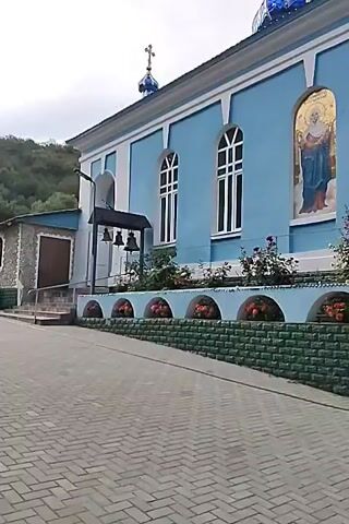 The video pans across the exterior of a blue and white building, which appears to be a church or monastery. In front of the building, a paved walkway leads to a large, dark cross adorned with flowers. Several flags are visible near the cross. To the left of the building is another smaller, stone structure with windows. In the background, a lush green hillside rises, dotted with trees. The sky is overcast. A few people are visible walking in the distance near the cross and to the left of the frame. Bells are hanging near the entrance of the main building. The overall impression is of a religious site in a natural, hilly environment.