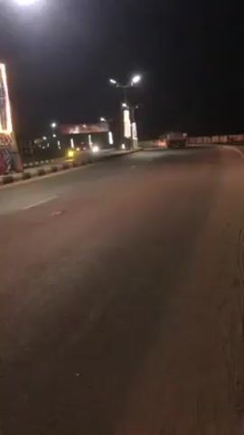 The video depicts a night-time street scene. Several cars and a motorcycle pass by, traveling away from the camera's perspective. Streetlights illuminate the road and decorative columns lining the roadside. A large sign is visible in the distance over the road. The road appears to be a multi-lane highway or major thoroughfare. The video is handheld and slightly shaky.