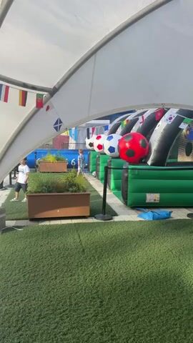 A person wearing a blue shirt and white shorts runs on a green grassy area under a white tent. The tent has various national flags hanging from its edge. Behind the person, there is an inflatable obstacle course with large soccer balls. In the background, people are seated and a soccer game is visible on a large screen or field. There are also potted plants on the grassy area.