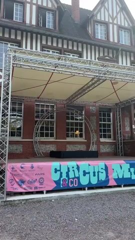 This video shows an outdoor stage set up in front of a large, Tudor-style building. The stage is covered by a metal truss structure with a fabric canopy. A banner across the front of the stage reads "CIRCUS & CO MICROFEST". On the stage, there is a black rectangular object. Behind the stage, visible through the windows of the building, are several objects, including what appears to be a rocking horse and a lamp. The ground in front of the stage is covered in gravel. The video pans slightly to the right, revealing more of the building and its facade.