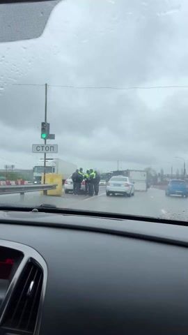 The video, filmed from inside a car, captures a road incident on an overcast and rainy day in Rogachevka, Russia. The dashboard of the filming vehicle is visible in the foreground, showing a digital display with "8°C" and raindrops clinging to the windshield, which is intermittently cleared by a wiper.

The main scene unfolds on a multi-lane road where a white refrigerator truck or trailer has overturned, lying on its side and obstructing traffic. Several individuals, including three people in what appear to be police or traffic officer uniforms (dark jackets with bright green-yellow reflective vests), are gathered near the overturned truck. One person in civilian clothing is also visible interacting with the officers.

A white police sedan, equipped with flashing blue and red lights on its roof, is parked nearby, facing away from the camera. Its license plate is partially visible, showing "M 0502". Cyrillic text, consistent with Russian police vehicles, is visible on its side. To the left of the scene, a "СТОП" (STOP) sign is clearly visible on a pole alongside traffic lights and some yellow and white barriers.

Other vehicles are present on the road: a large white truck is seen in the distance, and a blue car is driving away from the scene on the right. The overall atmosphere suggests a serious traffic incident requiring police intervention, with traffic moving slowly past the disruption on the wet road.