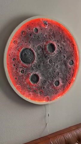 This media file features a close-up, slightly blurry view of a circular, illuminated art piece resembling the moon. The surface of the "moon" is textured with craters of varying sizes, painted in dark gray and black tones. A vibrant red light emanates from behind the outer edge of the piece, casting a warm glow and highlighting the lunar landscape. The artwork is mounted on a plain, light-colored wall. A thin white power cord dangles from the bottom center of the artwork, suggesting it is an electronic installation. The overall impression is one of an otherworldly, possibly stylized, celestial body. No people are visible in the frame. The context provided states the location as Huntington Beach, USA, however, there are no visual cues within the media file to confirm or deny this location. The time of day and weather are also not discernible from the image.