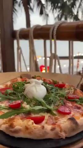 The video showcases a visually appealing pizza served on a wooden table with a beautiful beach view in the background. The pizza is topped with a white burrata cheese ball, arugula, and sliced tomatoes. The camera pans up to reveal a bamboo railing adorned with rope and a picturesque view of the ocean, palm trees, and other diners enjoying the beachside setting.