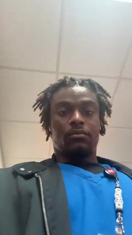A close-up, low-angle shot shows a person with dark skin, facial hair, and dreadlocks. The person has a nose ring. They are wearing a blue scrub top and a black jacket with a zipper.  A red and white ID badge hangs from their neckline. The person appears to be in a hallway with white tile ceiling and fluorescent lights.  The person moves slowly down the hallway, keeping the camera focused on their face. Blue and white signage are visible on the wall.  The overall impression is that the person is recording a selfie video while walking through a medical facility.