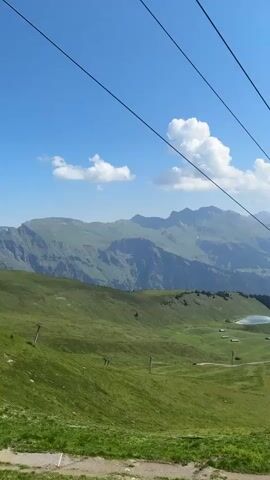 A cable car moves up a mountain on a sunny day. The cable car is dark gray and the cables are black. The mountain is covered in green grass and there are some trees scattered on the slopes. In the distance, there are more mountains with snow-capped peaks. The sky is blue with a few white clouds. There are a few people walking on a path in the foreground.