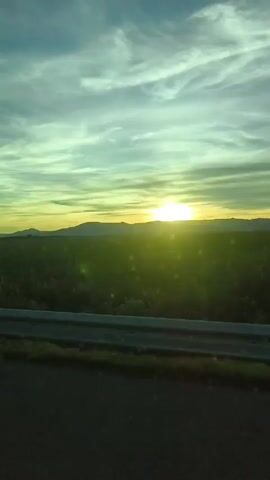 The video shows a landscape view taken from a moving vehicle. The camera is positioned low, capturing a stretch of asphalt road bordered by a gray metal guardrail. Beyond the guardrail, a vast green field extends toward the horizon, where a range of hills or low mountains is silhouetted against the bright light of the setting sun. The sky is a blend of pale blues, greens, and yellows, with streaks of clouds stretching across it. The overall lighting is bright due to the sun, and there's a noticeable lens flare effect from the sun. There are no people visible in the frame.