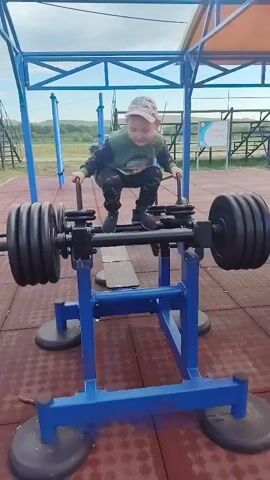 A young boy in a camo outfit and a cap is on a blue weight machine with heavy plates on both ends. He is squatting and holding onto handles. The machine appears to be outdoors on a rubberized surface. The boy moves up and down, as if he is using the machine to exercise. The background shows trees and a fence. The sky is cloudy.